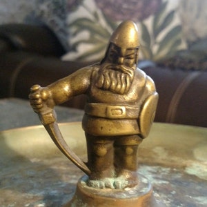 May include: A gold metal figurine of a warrior holding a sword and shield. The figurine is standing on a round base.