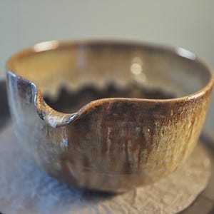 Puede incluir: Un cuenco de cerámica marrón con un borde ondulado y un esmalte moteado. El cuenco está sobre una superficie oscura.