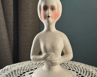 May include: A bisque doll head and torso, painted with delicate features. The doll has blue eyes, blushed cheeks, and a red mouth. The arms are crossed at the chest. The doll is displayed on a white lace tablecloth.