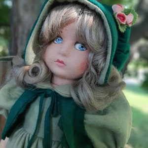 May include: A handmade doll with blonde hair, blue eyes, and a green and white dress. The doll is wearing a green hooded cloak with a floral trim.