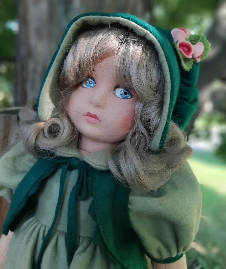 May include: A handmade doll with blonde hair, blue eyes, and a green and white dress. The doll is wearing a green hooded cloak with a floral trim.