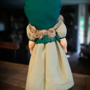 May include: A doll wearing a green dress with a white skirt and a green hat. The doll has blonde hair and is standing on a white stand.