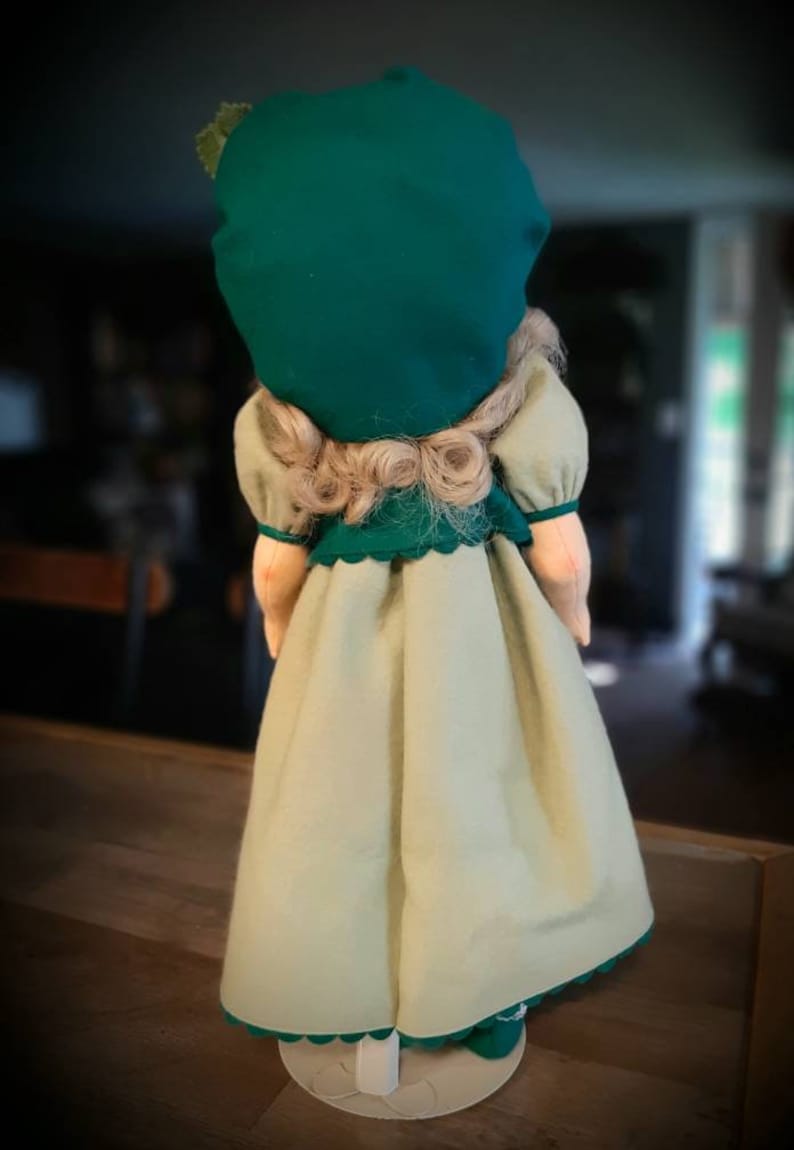 May include: A doll wearing a green dress with a white skirt and a green hat. The doll has blonde hair and is standing on a white stand.