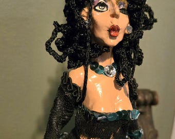 May include: A decorative figurine featuring a figure with curly black hair, a blue gemstone headpiece, and red lips. The figure wears a black lace top with long sleeves and a skirt adorned with shimmering teal sequins and a matching necklace.
