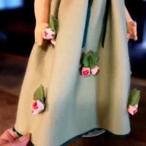 May include: A green dress with a scalloped hem and pink felt flowers. The dress has a green sash and a green collar.