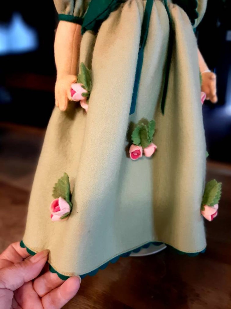 May include: A green dress with a scalloped hem and pink felt flowers. The dress has a green sash and a green collar.