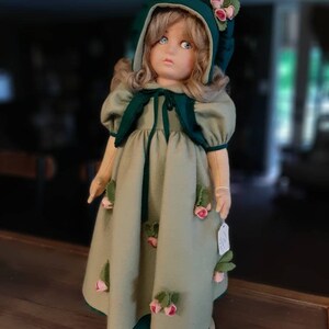 May include: A vintage porcelain doll wearing a green dress with rose accents and a green hooded hat. The doll has blonde hair and blue eyes.