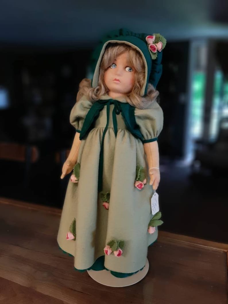 May include: A vintage porcelain doll wearing a green dress with rose accents and a green hooded hat. The doll has blonde hair and blue eyes.