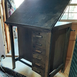 以下が含まれることがあります： 傾斜した天板と複数の引き出しが付いた、ヴィンテージの木製製図台。テーブルは濃い茶色で、使い込まれた仕上げになっています。