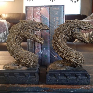 Puede incluir: Un par de sujetalibros de color dorado con forma de dragón con la boca abierta. Los sujetalibros están sobre una superficie de madera y sostienen un conjunto de libros de Harry Potter.