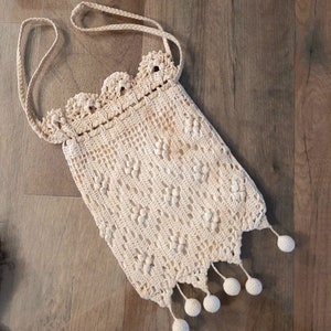 May include: A crocheted cream-colored bag with a long strap and a scalloped top. The bag has a fringe of white pom-poms.
