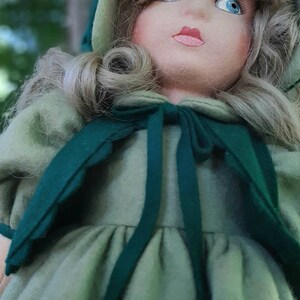 May include: A vintage doll with blonde hair wearing a green dress and a green hooded cape. The doll has blue eyes and a painted smile.