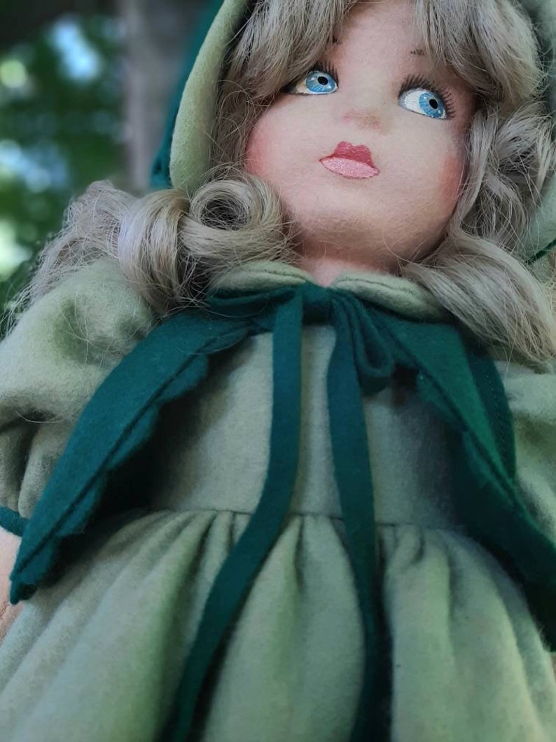 May include: A vintage doll with blonde hair wearing a green dress and a green hooded cape. The doll has blue eyes and a painted smile.
