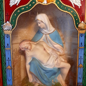 Può includere: Un'opera d'arte religiosa incorniciata che raffigura una donna in vesti blu e bianche che tiene un uomo tra le braccia. L'opera è su uno sfondo dorato e verde con un bordo rosso. Il testo "Blessed Be Thy Holy Name" è in basso nella cornice.