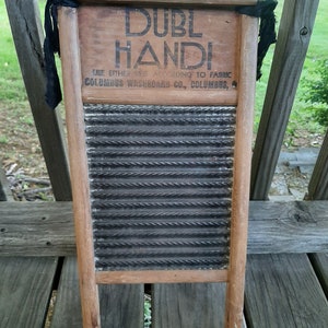 Op de afbeelding: Een vintage houten wasbord met een gegolfd oppervlak van zwart metaal. Het wasbord heeft een houten frame en twee houten handvatten. Op het wasbord staat de tekst "DUBL HANDI" en "COLUMBUS WASHBOARD CO., COLUMBUS, O.".