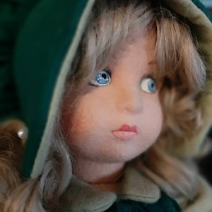 May include: A porcelain doll with blonde hair wearing a green hooded dress with a white trim. The doll has blue eyes and a painted smile.