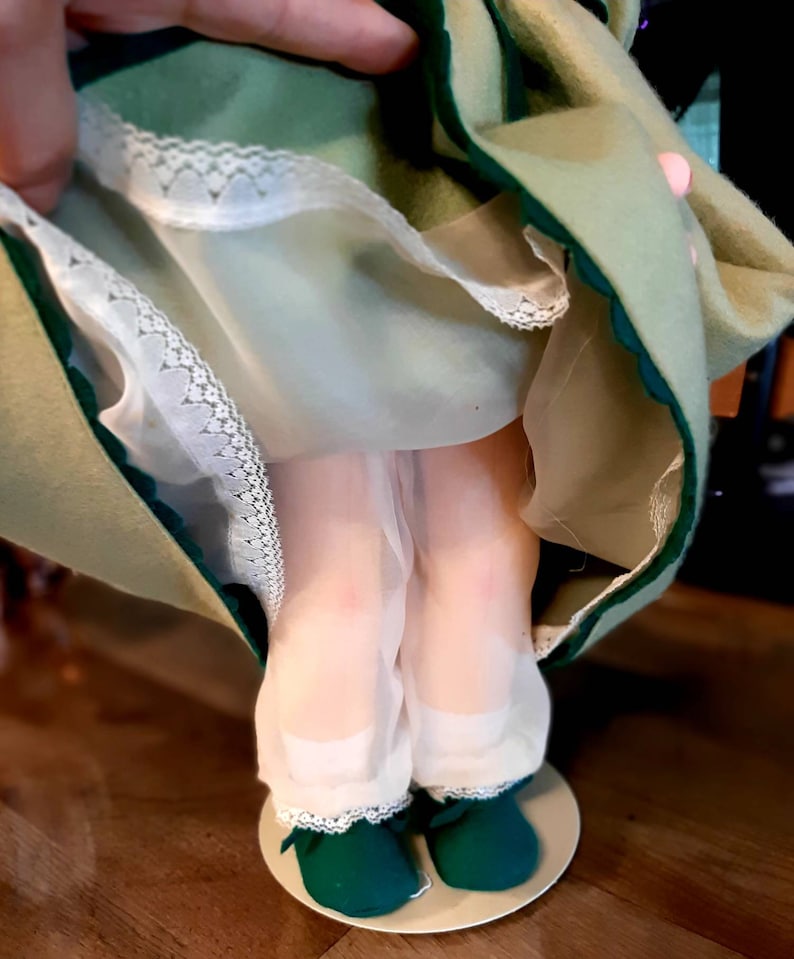 May include: A vintage doll wearing a green dress with white lace trim and green shoes. The doll is standing on a white pedestal.