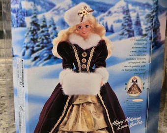 May include: A Barbie doll in a box with a winter holiday theme. The doll is wearing a burgundy velvet dress with gold accents and a white fur trim. The box has a winter scene background with the text "HAPPY HOLIDAYS Barbie DOLL".