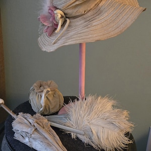 Puede incluir: Un sombrero vintage de ala ancha, adornado con plumas y una flor rosa, se exhibe sobre un soporte rosa. Un parasol abierto, una pequeña bolsa con cordón y un abanico de plumas completan la estética clásica y antigua.