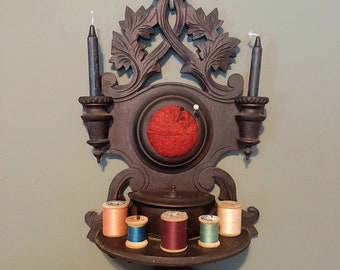 Könnte beinhalten: Ein dunkelbraunes, an der Wand montiertes Nadelkissen mit einem roten Samtmittelteil und einem Regal darunter zur Aufbewahrung von Garnrollen. Das Nadelkissen hat auf jeder Seite zwei Kerzenhalter.