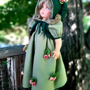 May include: A green felt doll with blonde hair wearing a green felt dress and a green felt hooded cloak. The doll has pink felt flowers on the dress and cloak.