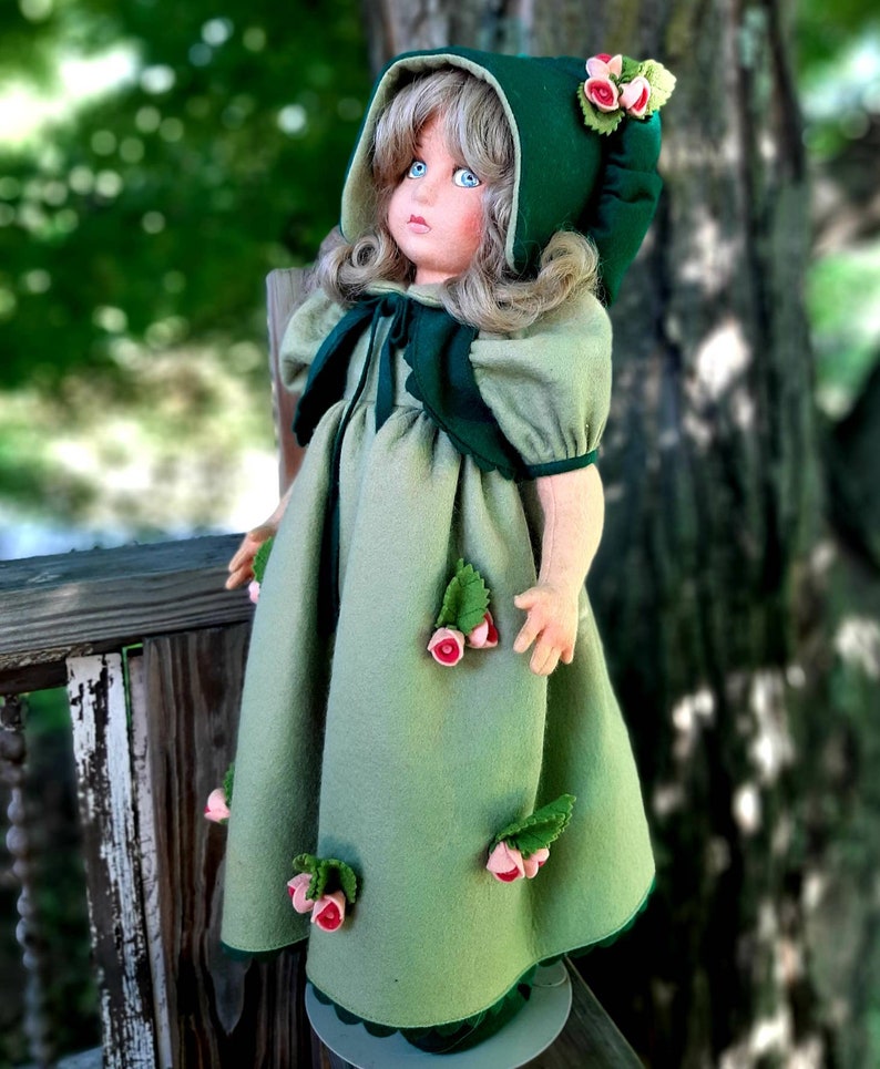 May include: A green felt doll with blonde hair wearing a green felt dress and a green felt hooded cloak. The doll has pink felt flowers on the dress and cloak.
