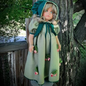 May include: A porcelain doll wearing a green dress with pink flower accents and a green bonnet. The doll has blonde hair and blue eyes.