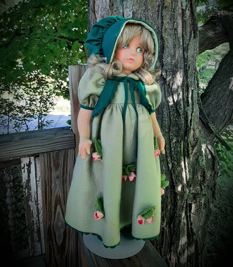 May include: A porcelain doll wearing a green dress with pink flower accents and a green bonnet. The doll has blonde hair and blue eyes.