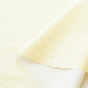 May include: Close-up of a pale yellow fabric with a white underside, partially rolled. Water droplets are scattered across the surface. The fabric has a smooth texture and a subtle sheen. The word "CHUNGAGE" is visible on the fabric.