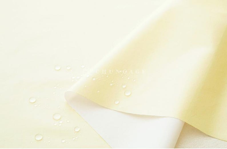 May include: Close-up of a pale yellow fabric with a white underside, partially rolled. Water droplets are scattered across the surface. The fabric has a smooth texture and a subtle sheen. The word "CHUNGAGE" is visible on the fabric.