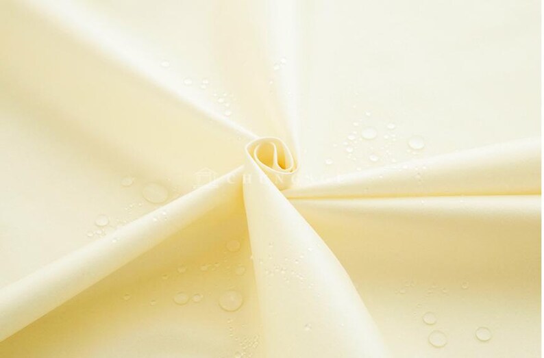 May include: Close-up of a pale yellow fabric with water droplets. The fabric is folded and rolled, creating a spiral shape. The material appears smooth and possibly water-resistant, with visible water beads.