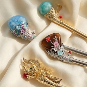 May include: Four ornate hair sticks with decorative tops. One features a golden dragon, another a brown stone with floral accents, and two with blue and green stone tops. The sticks are set against a cream-colored fabric.