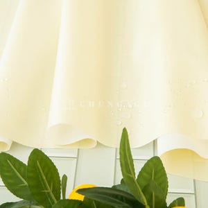 May include: A close-up of a pale yellow fabric with water droplets, possibly a tablecloth or a similar textile. The fabric is draped, with soft folds and a subtle sheen. Green leaves and yellow fruit are visible at the bottom.