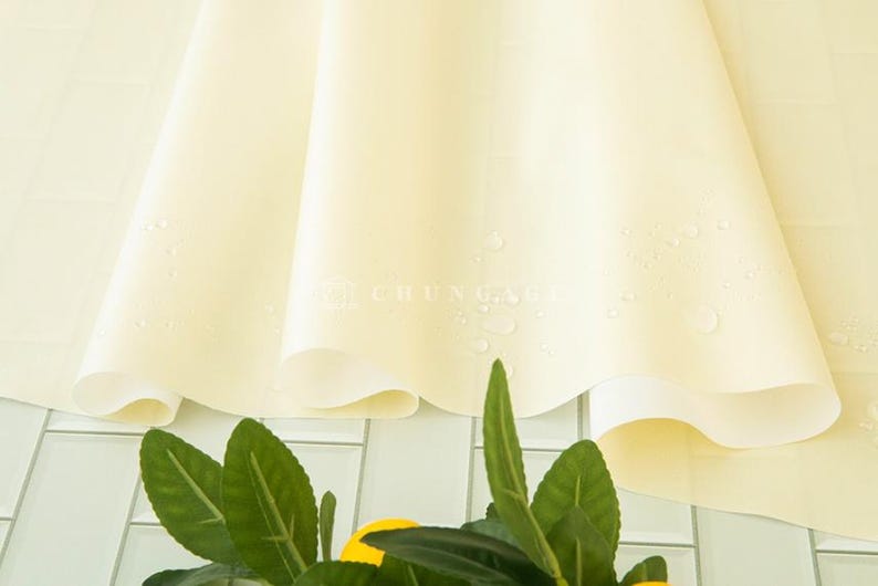 May include: A close-up of a pale yellow fabric with water droplets, possibly a tablecloth or a similar textile. The fabric is draped, with soft folds and a subtle sheen. Green leaves and yellow fruit are visible at the bottom.