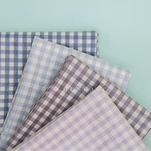 May include: A collection of folded fabric squares in various shades of blue, purple, and gray, all featuring a classic gingham pattern. The fabrics are arranged against a light teal background, creating a visually appealing display.