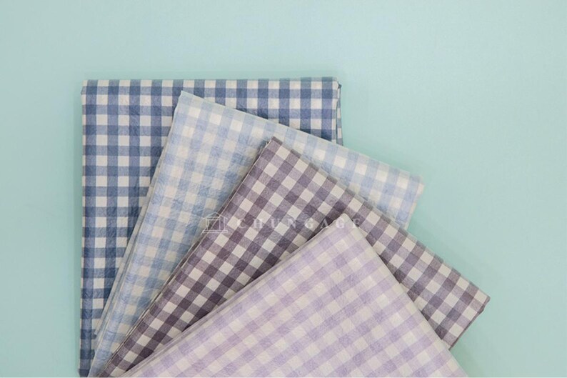 May include: A collection of folded fabric squares in various shades of blue, purple, and gray, all featuring a classic gingham pattern. The fabrics are arranged against a light teal background, creating a visually appealing display.