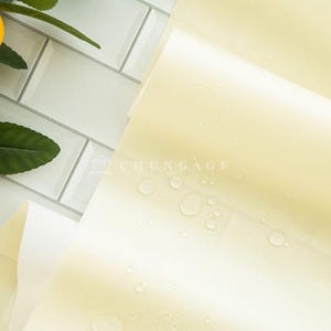 May include: A close-up shot of a stack of pale yellow paper with water droplets, next to a lemon and green leaves. The paper is slightly translucent, and the background is a white tile.