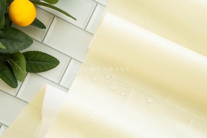 May include: A close-up shot of a stack of pale yellow paper with water droplets, next to a lemon and green leaves. The paper is slightly translucent, and the background is a white tile.