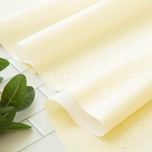 May include: A close-up shot of rolled, pale yellow fabric with water droplets, possibly a tablecloth or similar textile. A sprig of lemons with green leaves is in the corner, suggesting a fresh, natural theme.