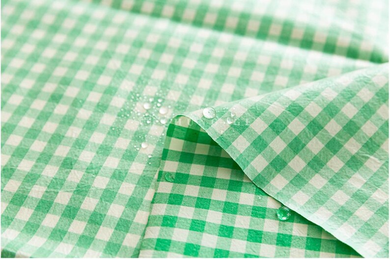 May include: Close-up of a green and white checkered fabric with water droplets on the surface. The fabric is folded, revealing the pattern and texture. The gingham pattern is a classic design.
