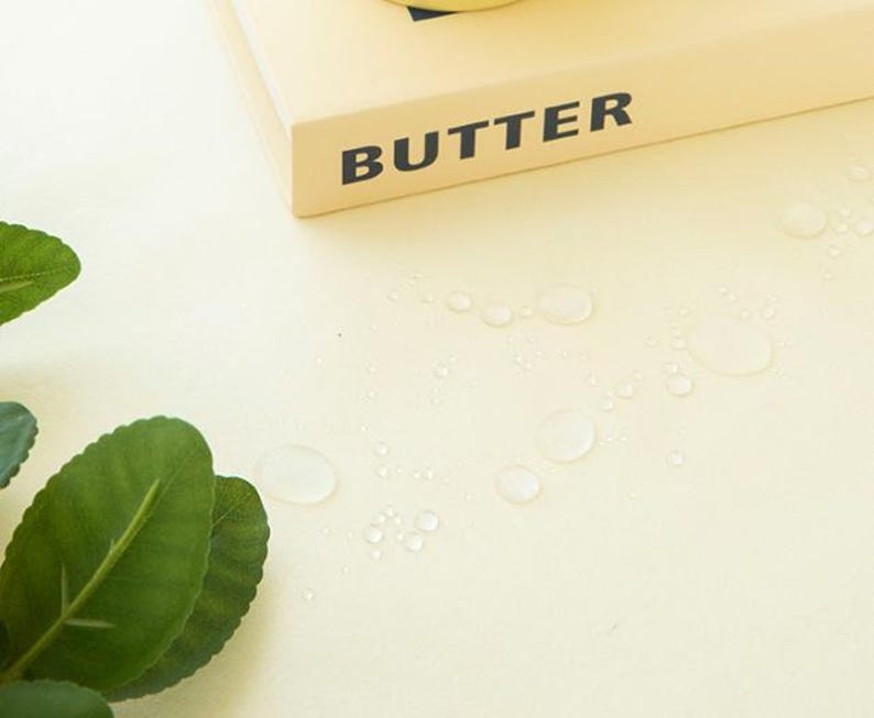 May include: A close-up shot of a light yellow surface with water droplets and green leaves. The word "BUTTER" is printed in black on a light yellow rectangular object.