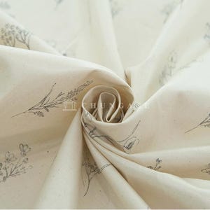 May include: Close-up of a cream-colored fabric with a delicate floral pattern printed in black. The fabric is folded and draped, showcasing its soft texture.