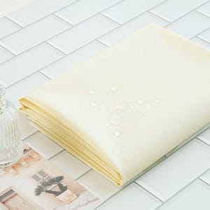 May include: A stack of folded, light yellow fabric with water droplets on top. A small, clear glass vase with a green plant and a picture of a building are next to the fabric. The items are on a white tiled surface.