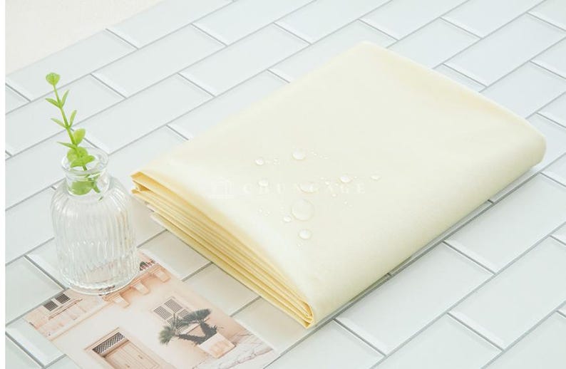 May include: A stack of folded, light yellow fabric with water droplets on top. A small, clear glass vase with a green plant and a picture of a building are next to the fabric. The items are on a white tiled surface.