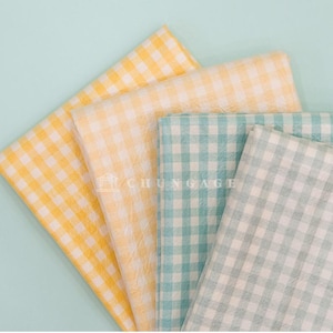 May include: An overhead shot displays four folded fabric squares in yellow, pale yellow, teal, and light green, all with a classic gingham pattern. The fabrics are arranged on a light blue background, creating a visually appealing display.