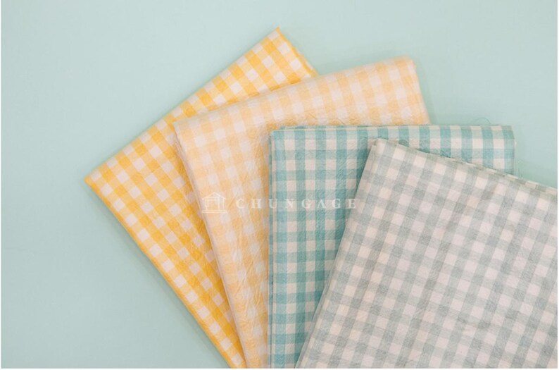 May include: An overhead shot displays four folded fabric squares in yellow, pale yellow, teal, and light green, all with a classic gingham pattern. The fabrics are arranged on a light blue background, creating a visually appealing display.