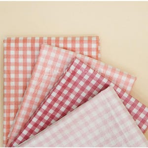 May include: A collection of folded fabric pieces in various shades of red and pink, featuring a classic gingham pattern. The fabrics are arranged on a light beige background, creating a visually appealing display.