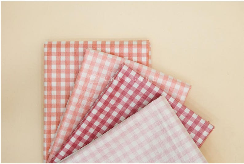 May include: A collection of folded fabric pieces in various shades of red and pink, featuring a classic gingham pattern. The fabrics are arranged on a light beige background, creating a visually appealing display.