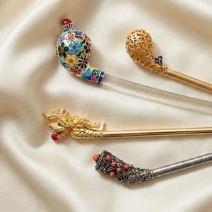 May include: Four ornate hair sticks with decorative tops. One features a floral design with red, blue, yellow, and green enamel. Another has a gold filigree design. A third has a gold dragon head. The last has a silver filigree design with red beads.