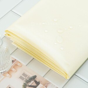 May include: A stack of folded, pale yellow fabric with water droplets on the surface. The fabric is neatly folded, revealing multiple layers. The background features a white tiled surface and a glass vase.